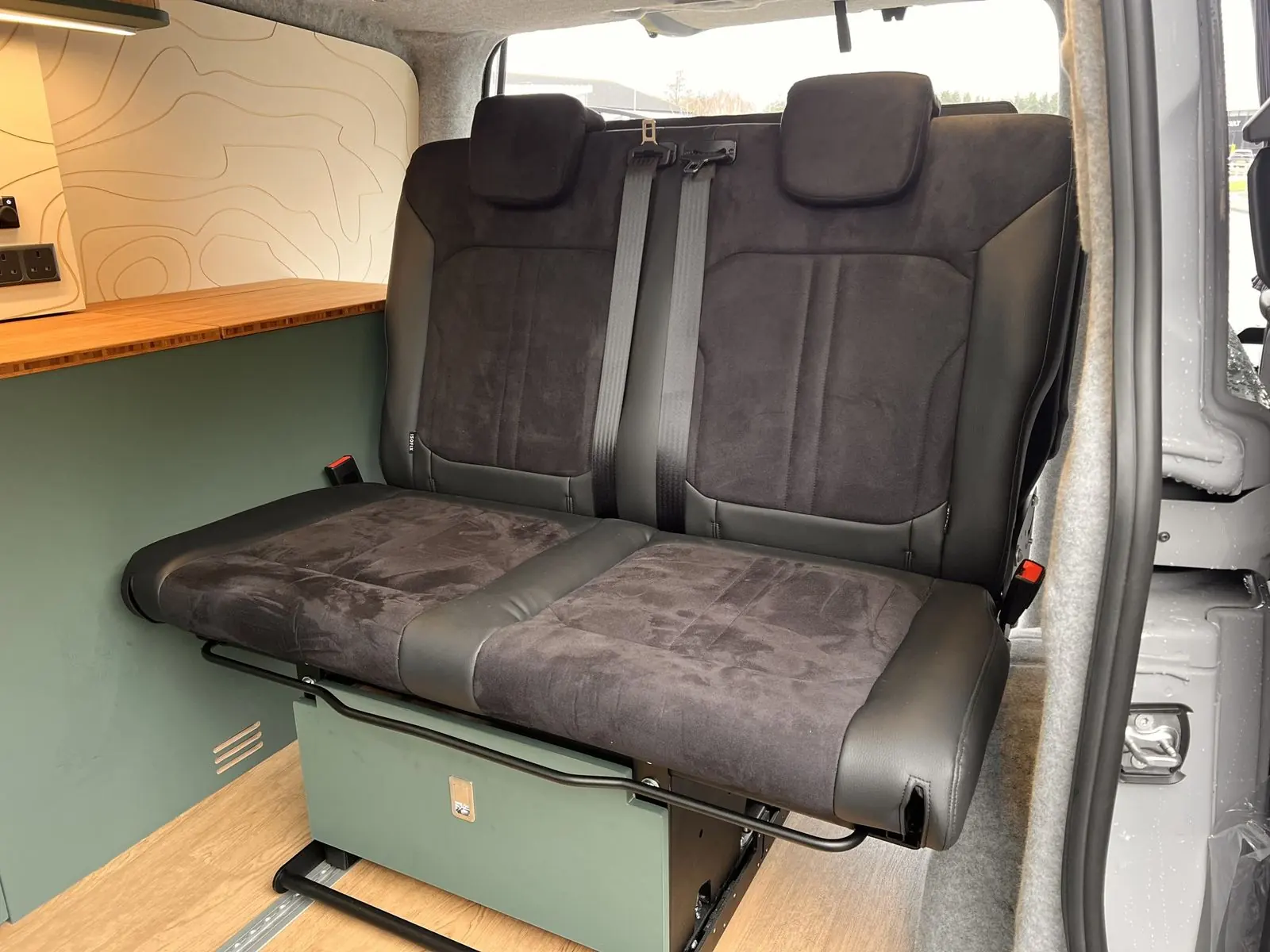 Two-seat rear bench in black suede-effect fabric with leather-look bolsters and black seat belts, on a dark green base.