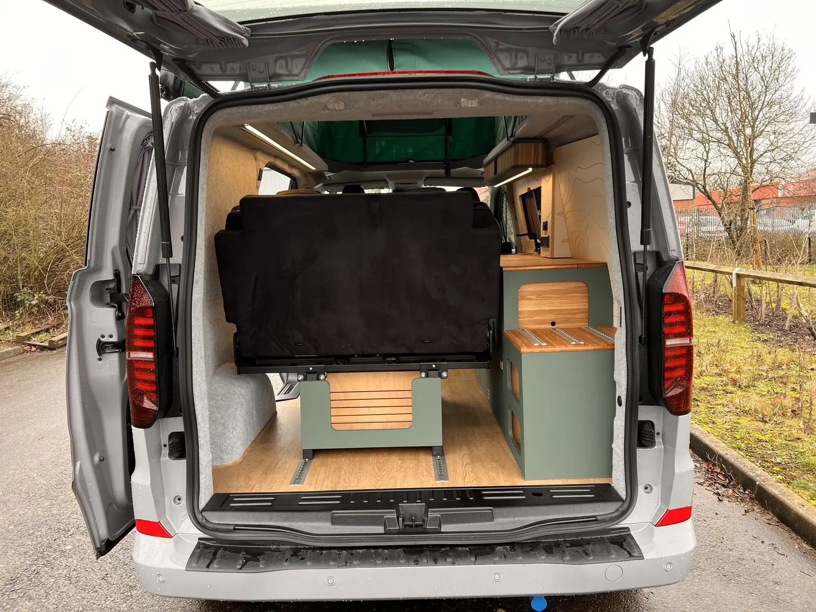 View in through the open tailgate showing the back of the rear seat, a dark green rear storage unit with a bamboo top and rails, a green seat base, oak-effect floor and the green pop-top canvas above.