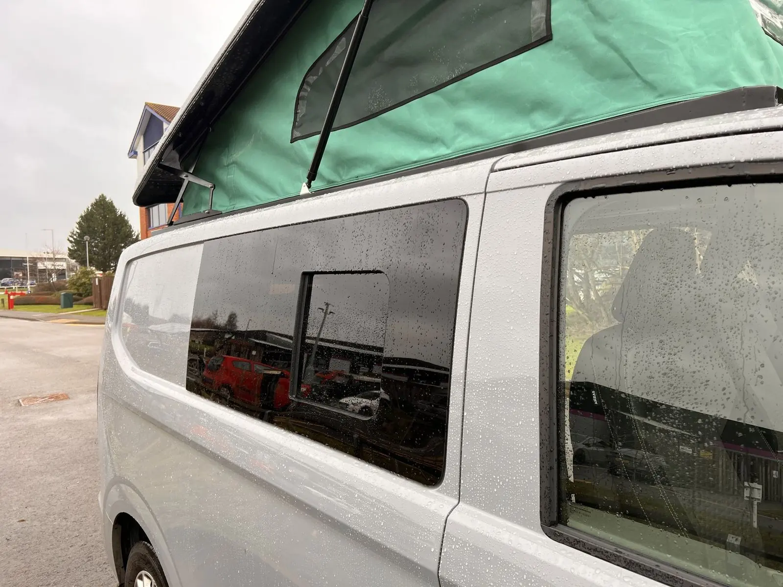Side of the light grey van with the raised pop-top showing green canvas with a mesh vent panel, above a tinted side window with a sliding pane.