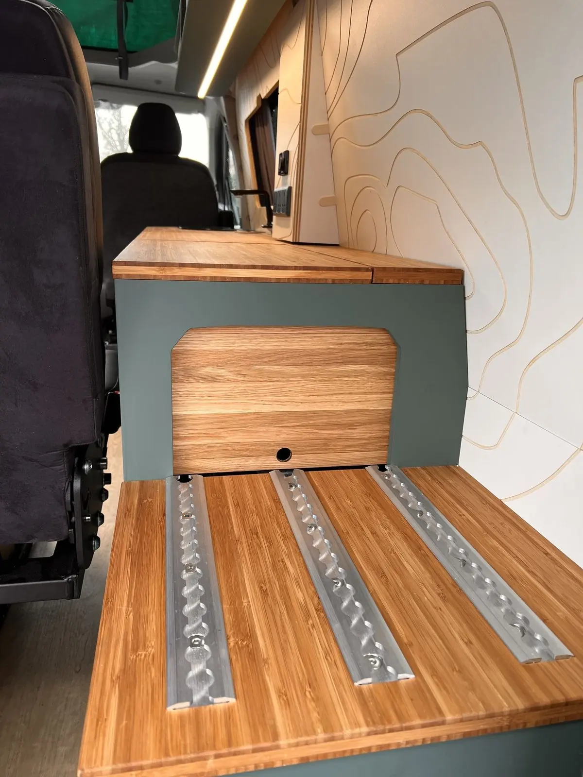 Rear storage unit in dark green with a bamboo top fitted with three aluminium rails and an oak tambour door, beside cream contour-line wall panels.