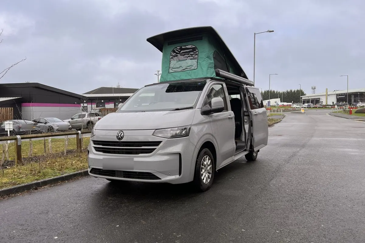Front three-quarter view of a light grey new-shape Volkswagen Transporter campervan on a wet road with its pop-top roof raised, showing green canvas, and the side door open.