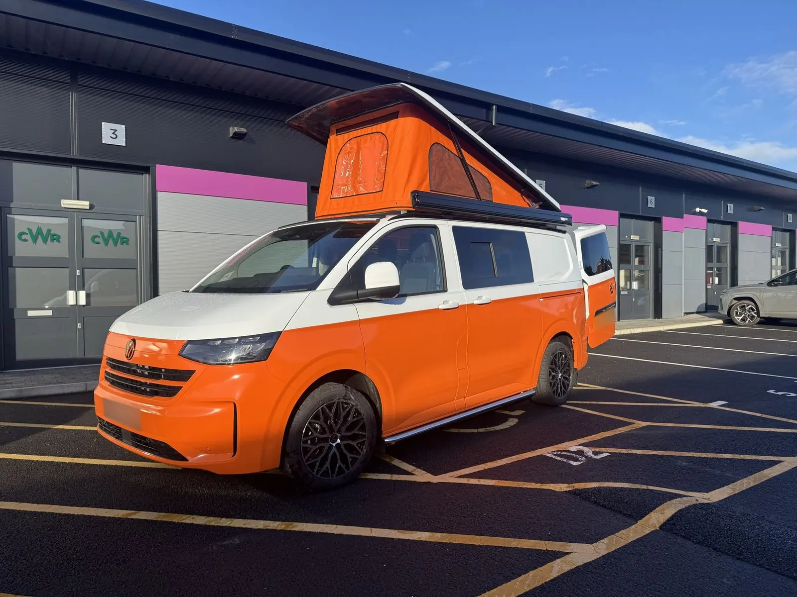 Front three-quarter view of a new-shape Volkswagen Transporter campervan in orange with a white upper half, orange pop-top roof raised, a black awning on the roof edge and black alloy wheels, parked outside a business unit.