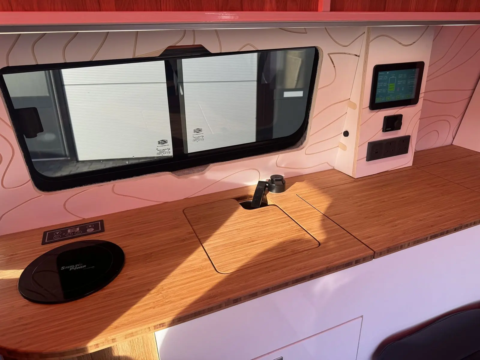 Bamboo worktop with a round black Sterling Power induction hob, a flush bamboo sink cover and black tap, below a sliding side window and a wall panel with a touchscreen, dial controller and sockets.