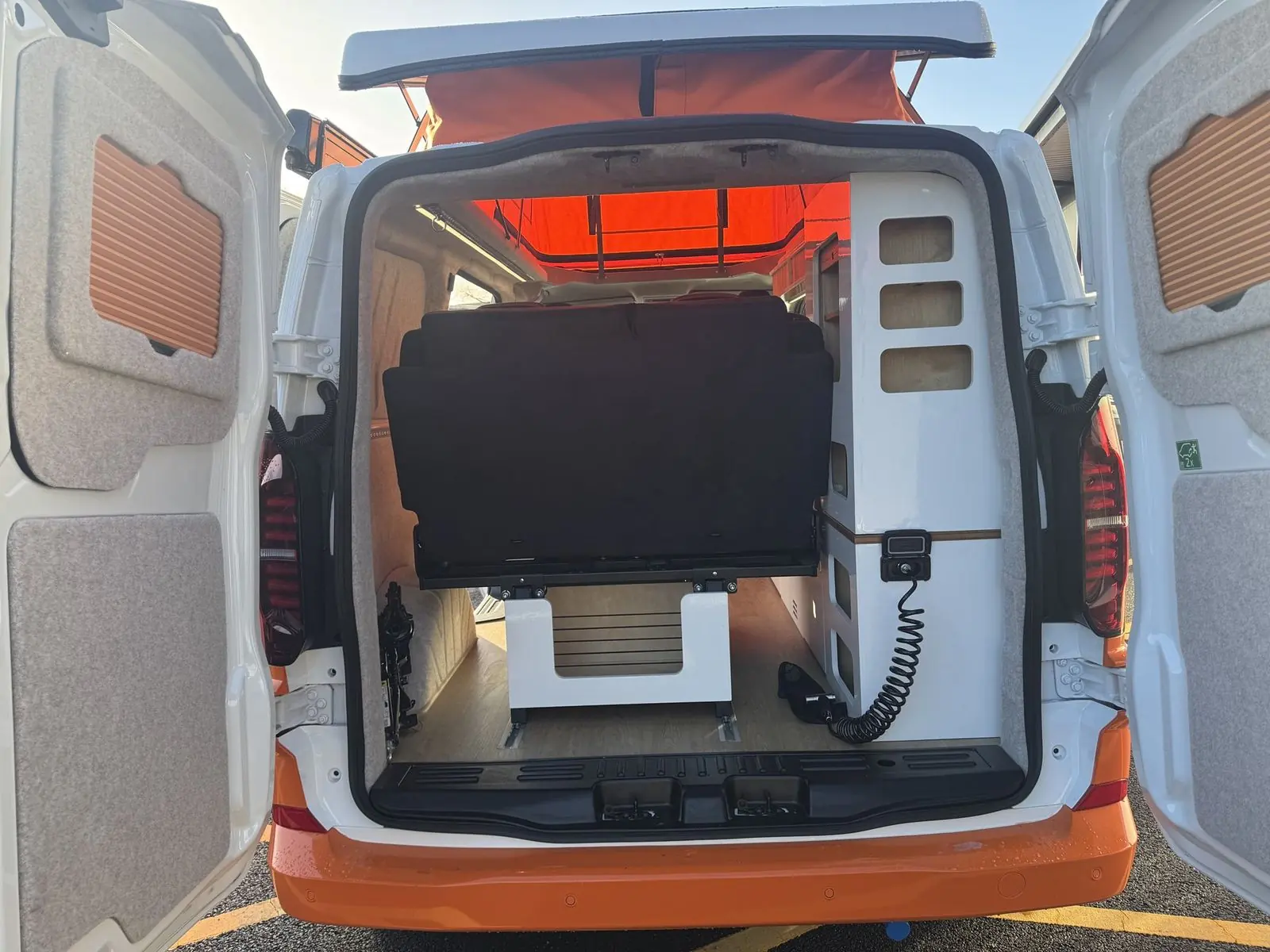 View in through the open rear barn doors showing the back of the dark rear seat, a tall white storage unit with rounded cubby holes, lined door panels with slatted wood inserts and the orange pop-top canvas above.