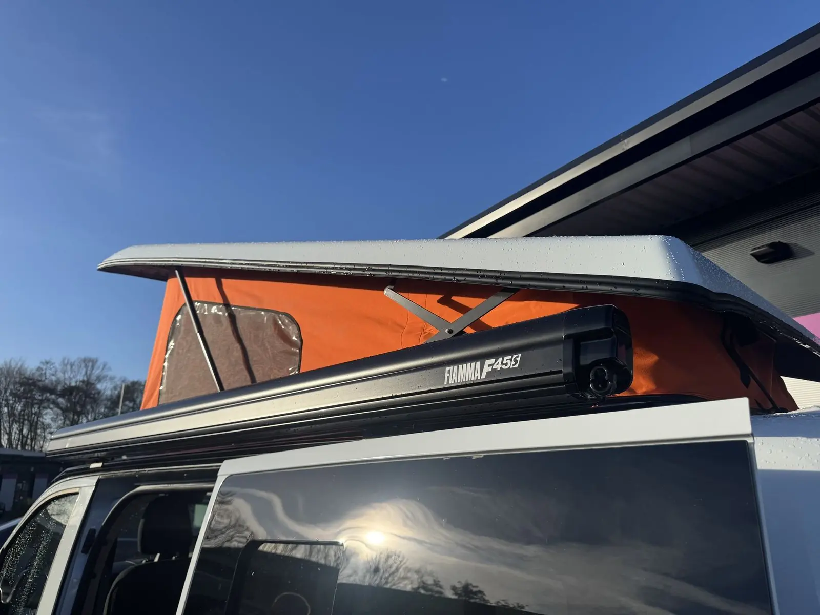 Raised pop-top roof with orange canvas and a clear window panel, with a black Fiamma F45s awning mounted along the roof edge.