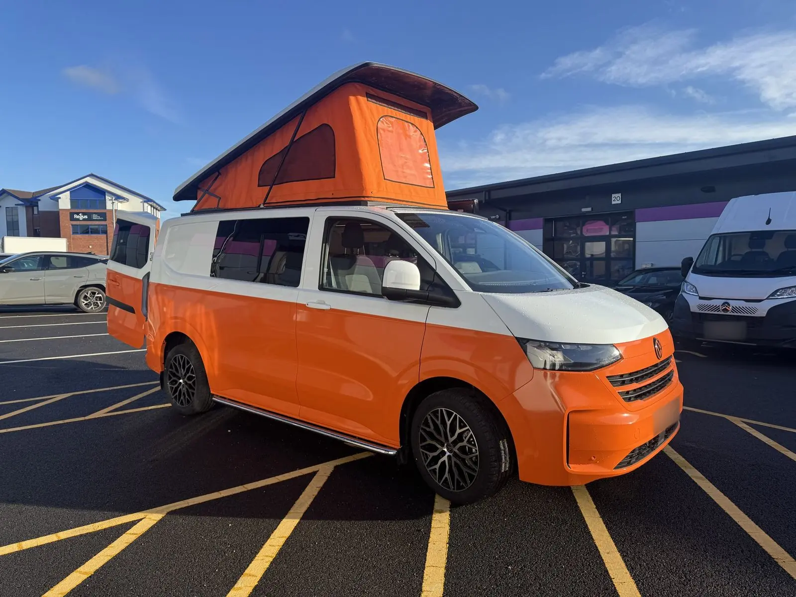 Side view of the orange and white campervan from the driver's side with the orange pop-top raised, black alloy wheels and chrome side bars.