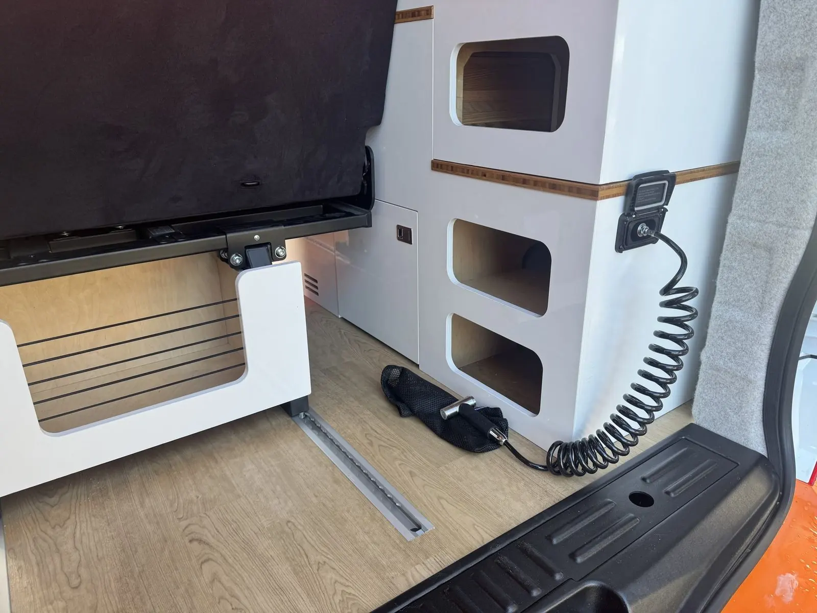 Rear load area with a white storage unit with rounded cubby holes and bamboo trim, a white seat base with a slatted plywood panel, a coiled cable on a wall socket, oak-effect floor and a floor rail.