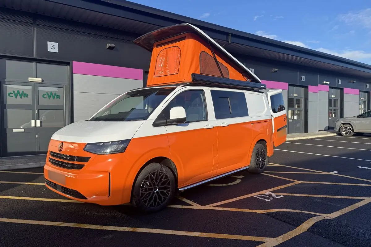 Front three-quarter view of a new-shape Volkswagen Transporter campervan in orange with a white upper half, orange pop-top roof raised, a black awning on the roof edge and black alloy wheels, parked outside a business unit.
