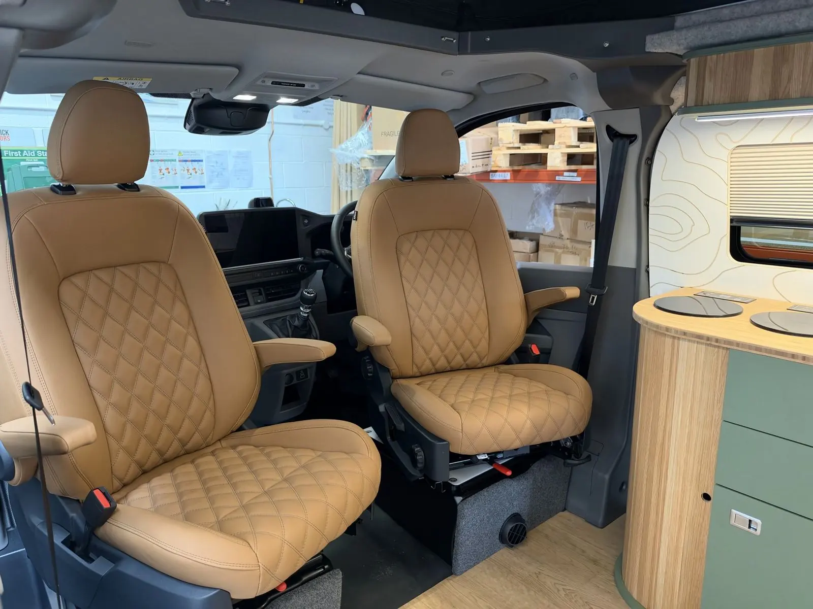 Two tan leather-look front seats with diamond-quilted centre panels and armrests, seen from the living area, beside a sage green kitchen unit with a slatted oak curved end and bamboo worktop with two round hobs.