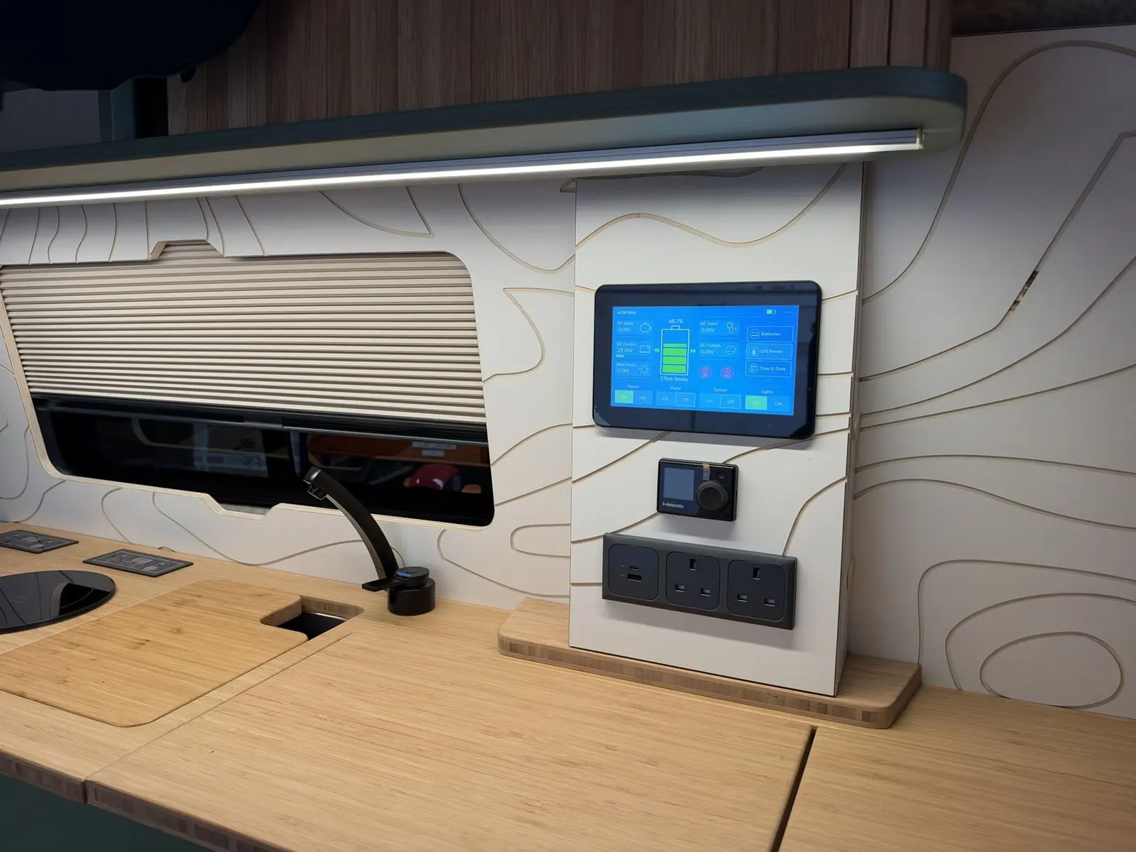 Kitchen wall with a bamboo worktop, black tap and part-covered sink, cream pleated window blind, and a white contour-line panel holding a touchscreen power display, Webasto controller and triple socket, under an overhead locker with LED strip.