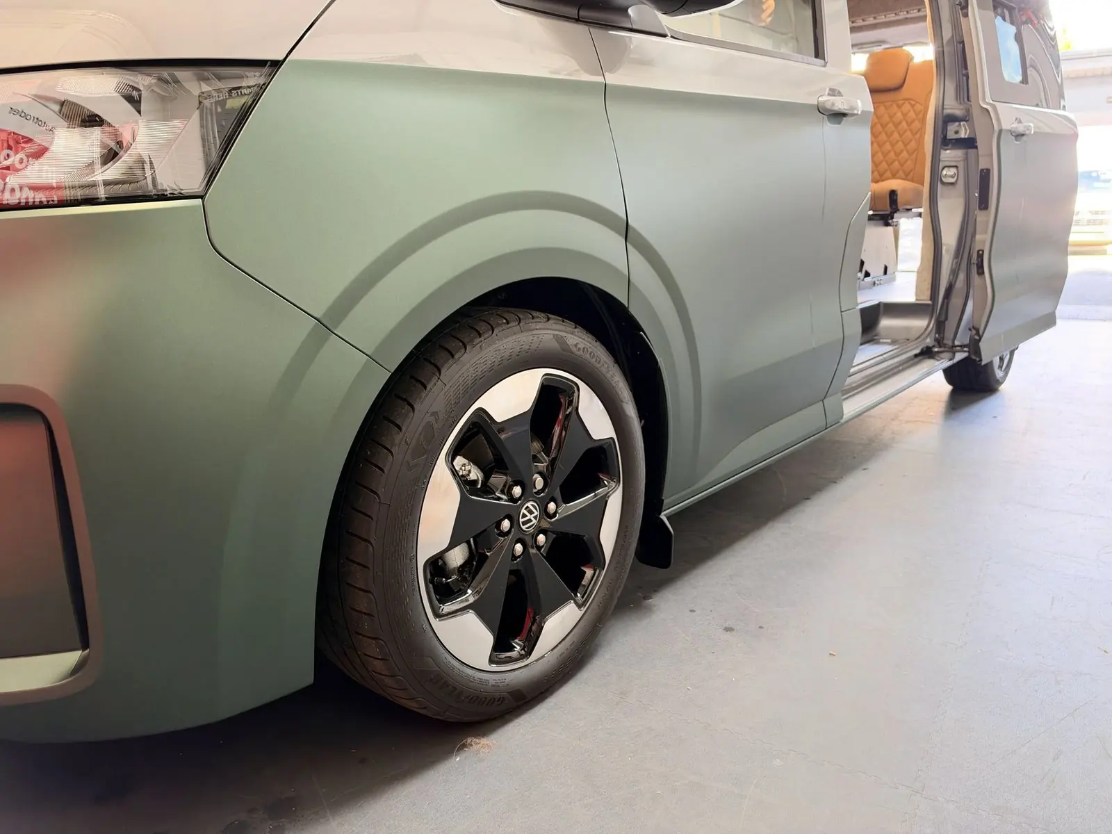 Low exterior view of the front wheel arch showing matt sage green lower bodywork, silver upper bodywork, a black and polished alloy wheel, and the sliding door open with a tan quilted seat visible inside.