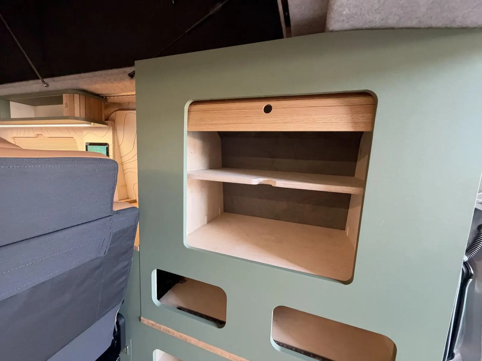 Sage green rear storage unit with a rounded cut-out revealing birch plywood shelves and an oak tambour-style door, with smaller open cubby holes below.
