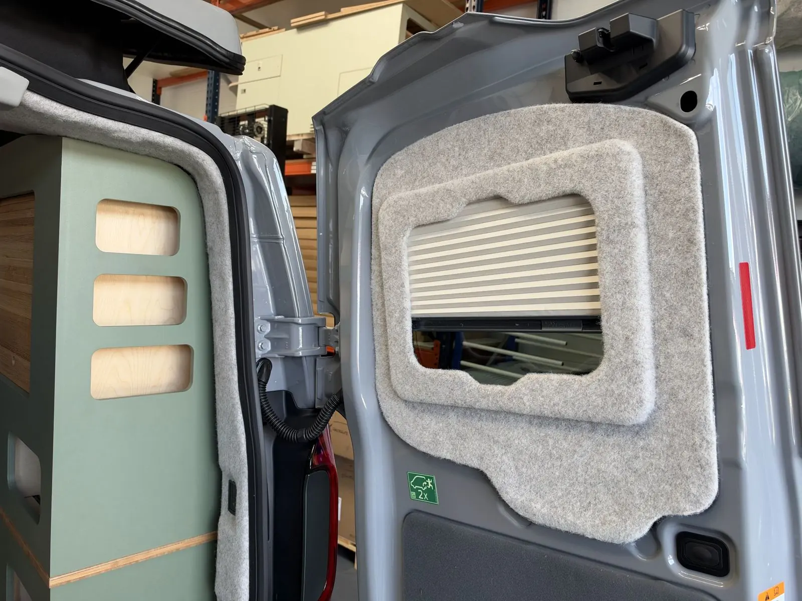 Open rear door lined with light grey carpet and fitted with a cream pleated blind over the window, next to the end of the sage green storage unit with three cut-out openings.