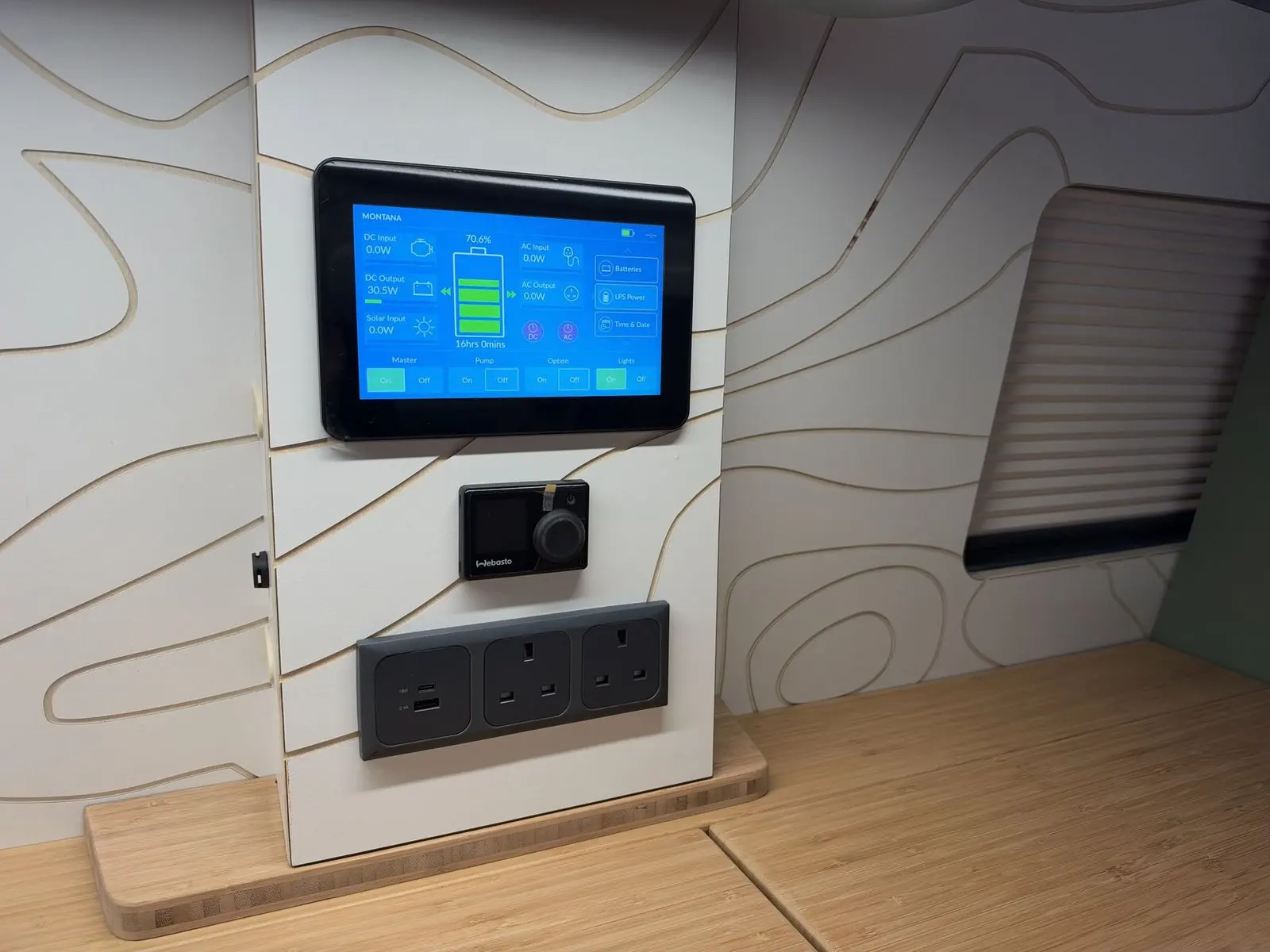 Close-up of the wall control panel: a touchscreen showing battery level and power inputs and outputs, a Webasto dial controller, and a grey triple socket with USB ports, mounted on a white contour-line panel.