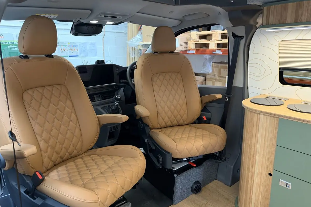 Two tan leather-look front seats with diamond-quilted centre panels and armrests, seen from the living area, beside a sage green kitchen unit with a slatted oak curved end and bamboo worktop with two round hobs.