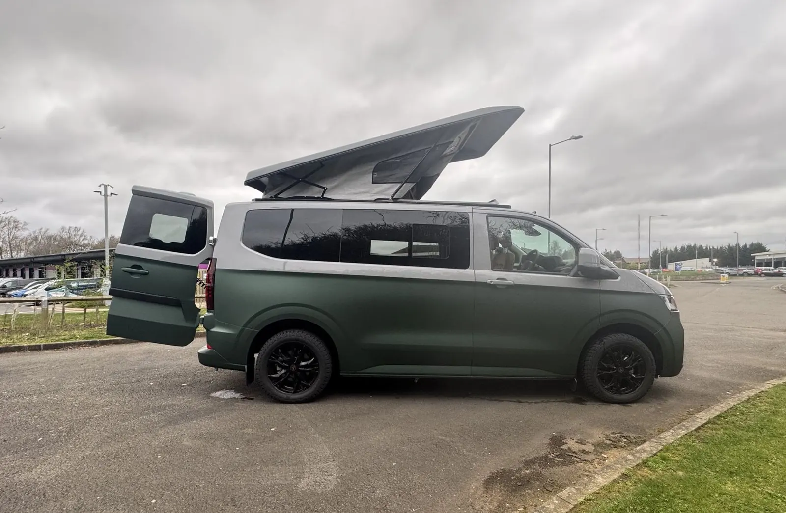 Side profile of a Volkswagen Transporter campervan in matt dark green with a silver upper half, black alloy wheels, pop-top roof raised and one rear door open, parked on tarmac under an overcast sky.