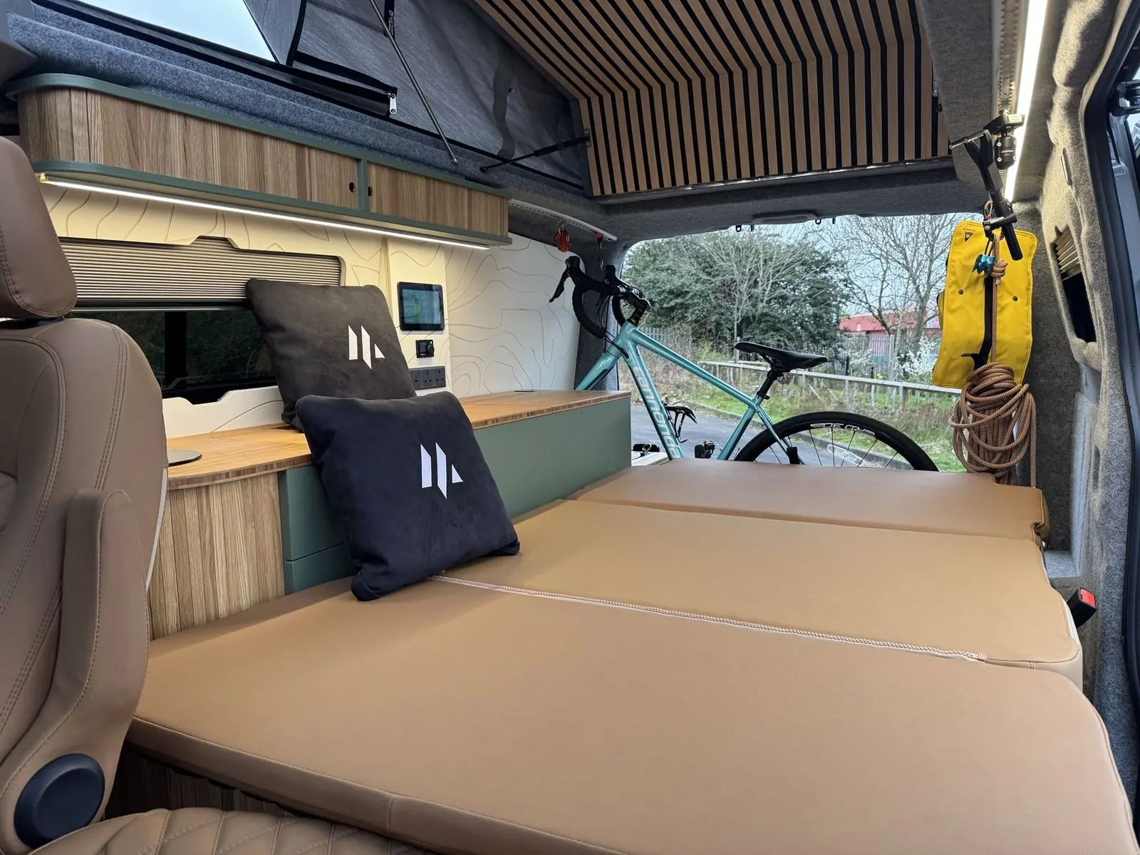 Interior with the rear seat folded flat into a tan bed with two black cushions, sage green kitchen unit with bamboo worktop, oak overhead locker, slatted wood roof panel, and a bicycle and climbing rope stowed at the rear.
