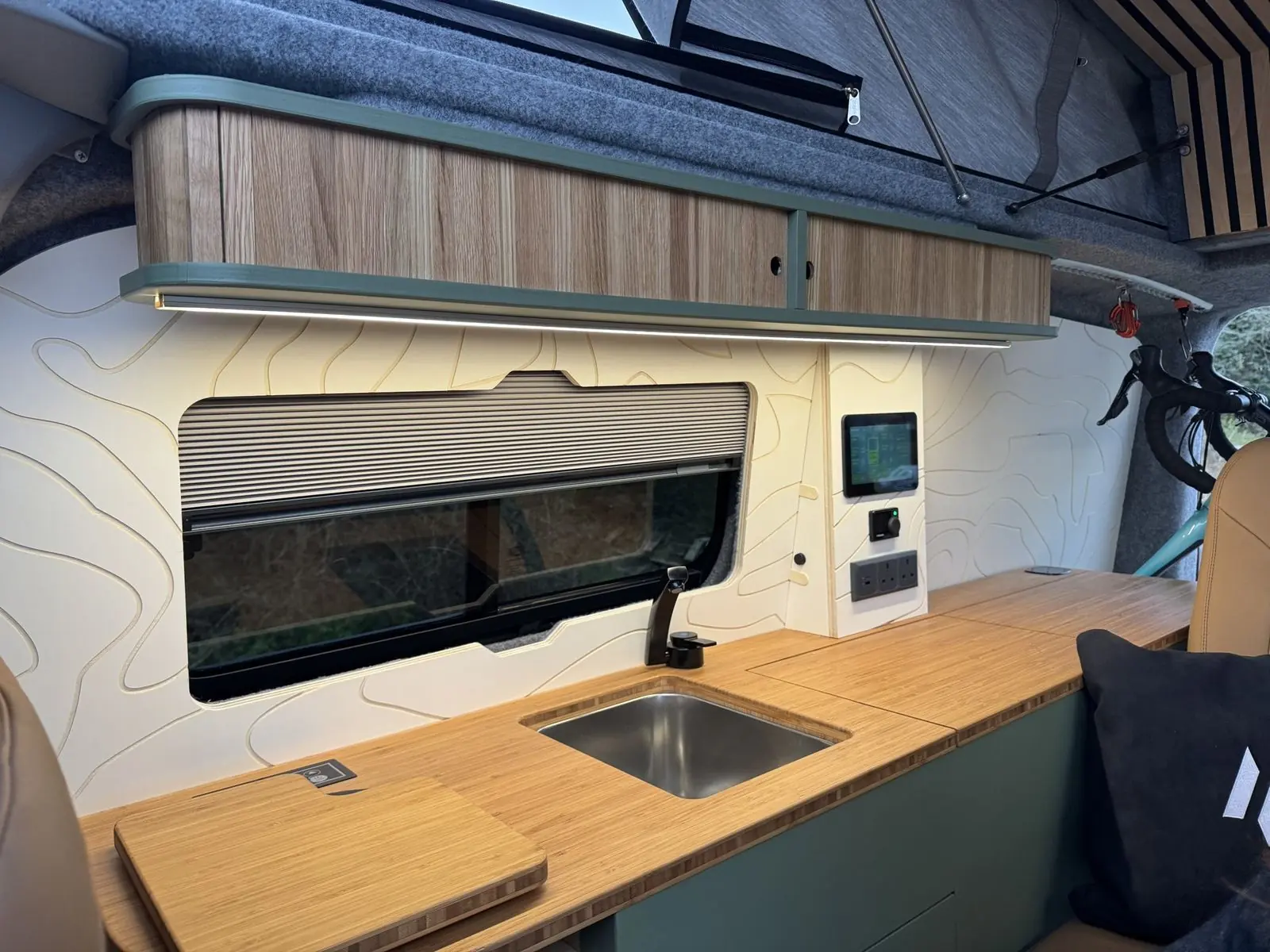 Kitchen wall with a stainless steel sink set into a bamboo worktop, black tap, pleated window blind, oak overhead locker with LED strip, and a touchscreen, dial controller and sockets on a white contour-line panel.