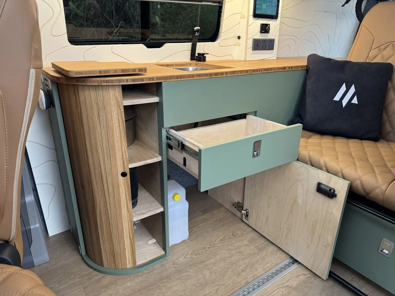 Sage green kitchen unit with a drawer and cupboard door open, a slatted oak curved end with open shelves, bamboo worktop, and a tan diamond-quilted rear seat with a black cushion.