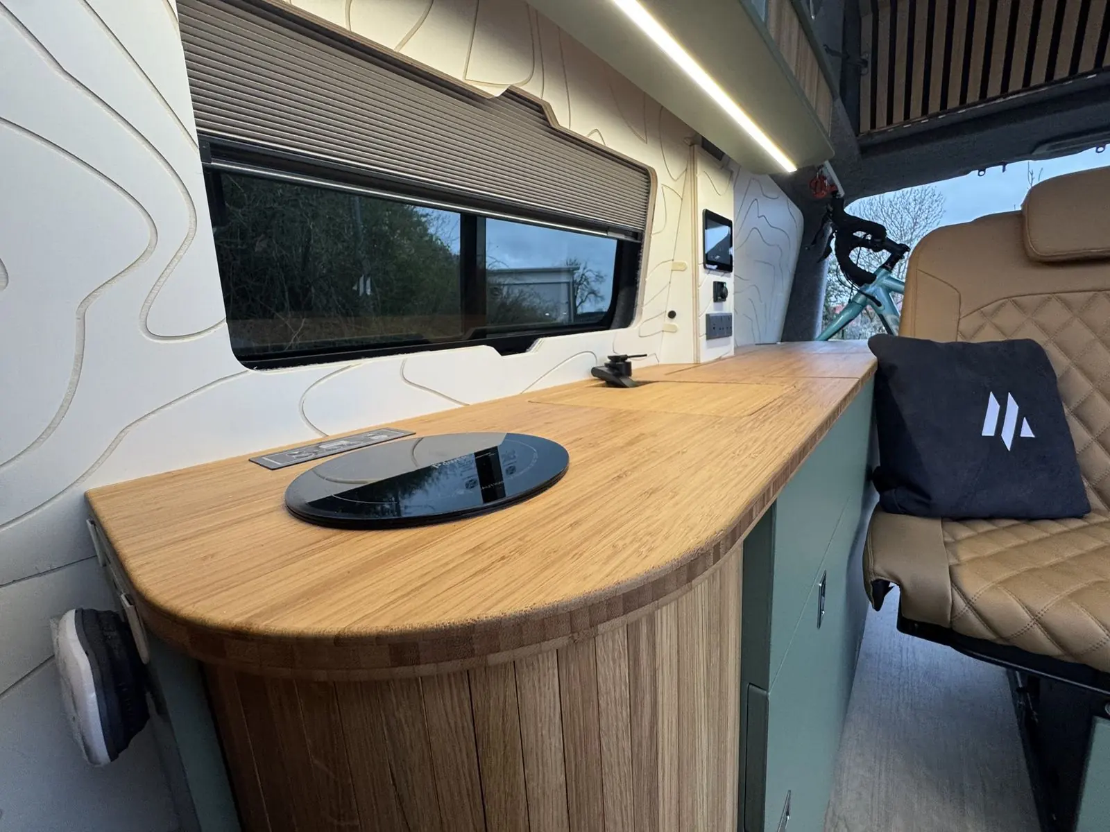 Bamboo worktop with a rounded end over slatted oak, a round black induction hob and a black tap, below a window with a pleated blind and a white contour-line wall panel.