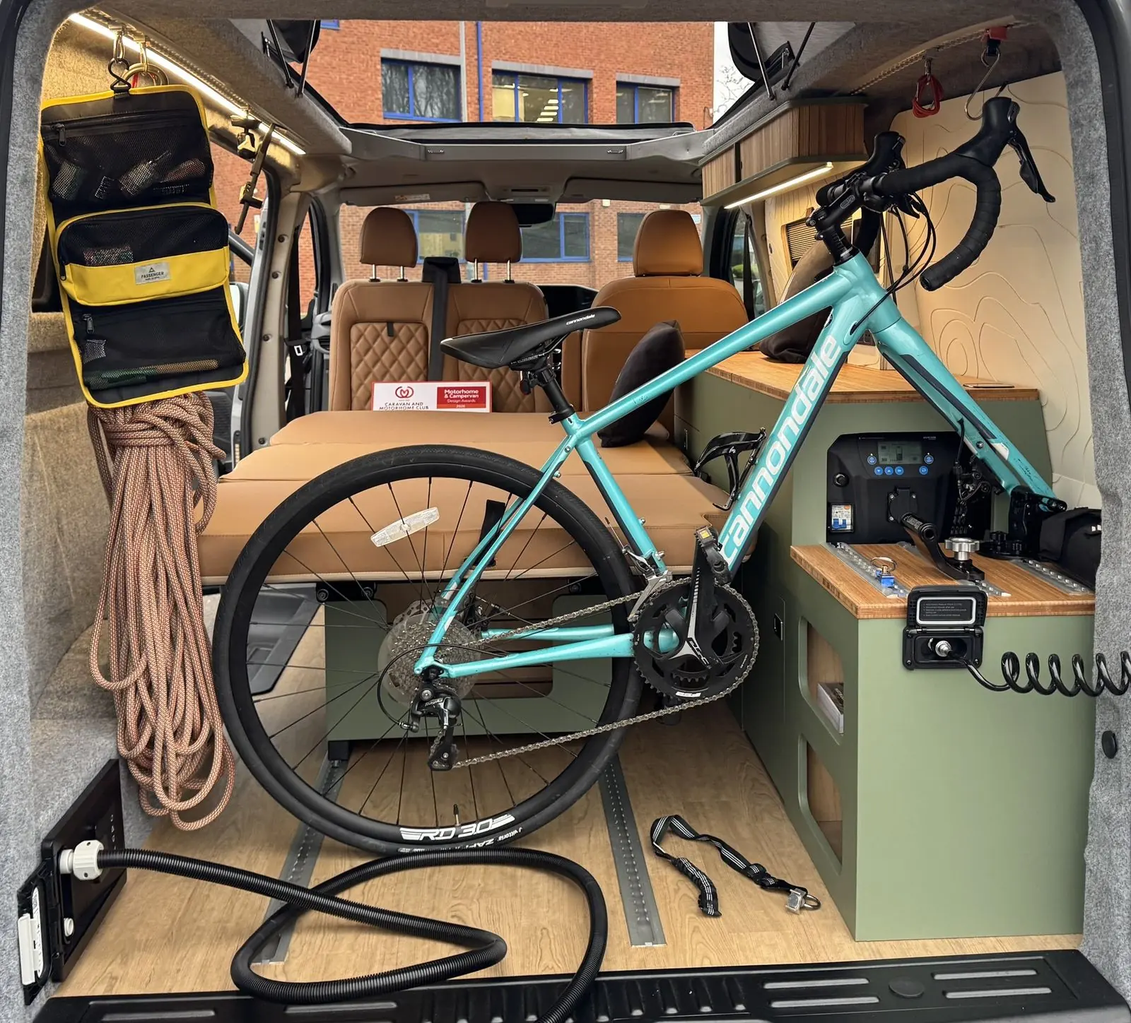 View in through the rear doors showing a turquoise road bike mounted upright beside a sage green storage unit with a built-in power unit, the bed laid flat, oak-effect floor with rails, and a rope and kit bag hanging from the ceiling rail.