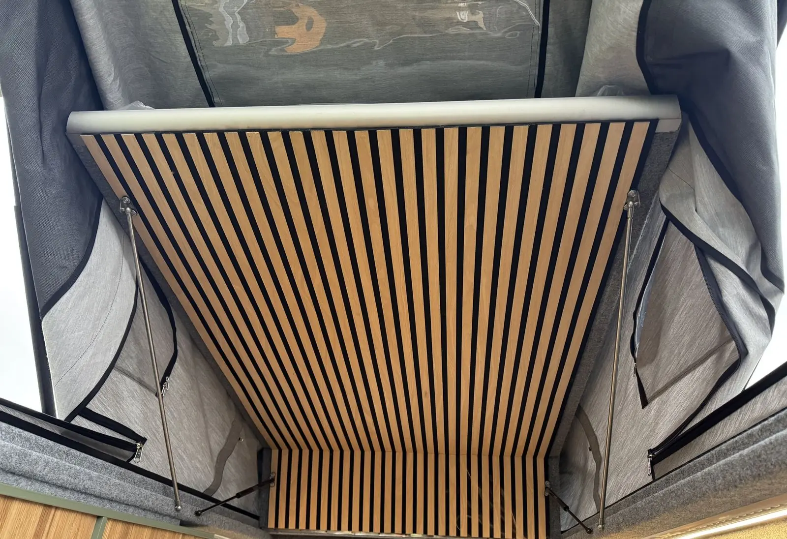 Looking up into the raised pop-top roof showing a slatted wood-and-black ceiling panel, light grey canvas sides with zipped window panels, and gas struts.