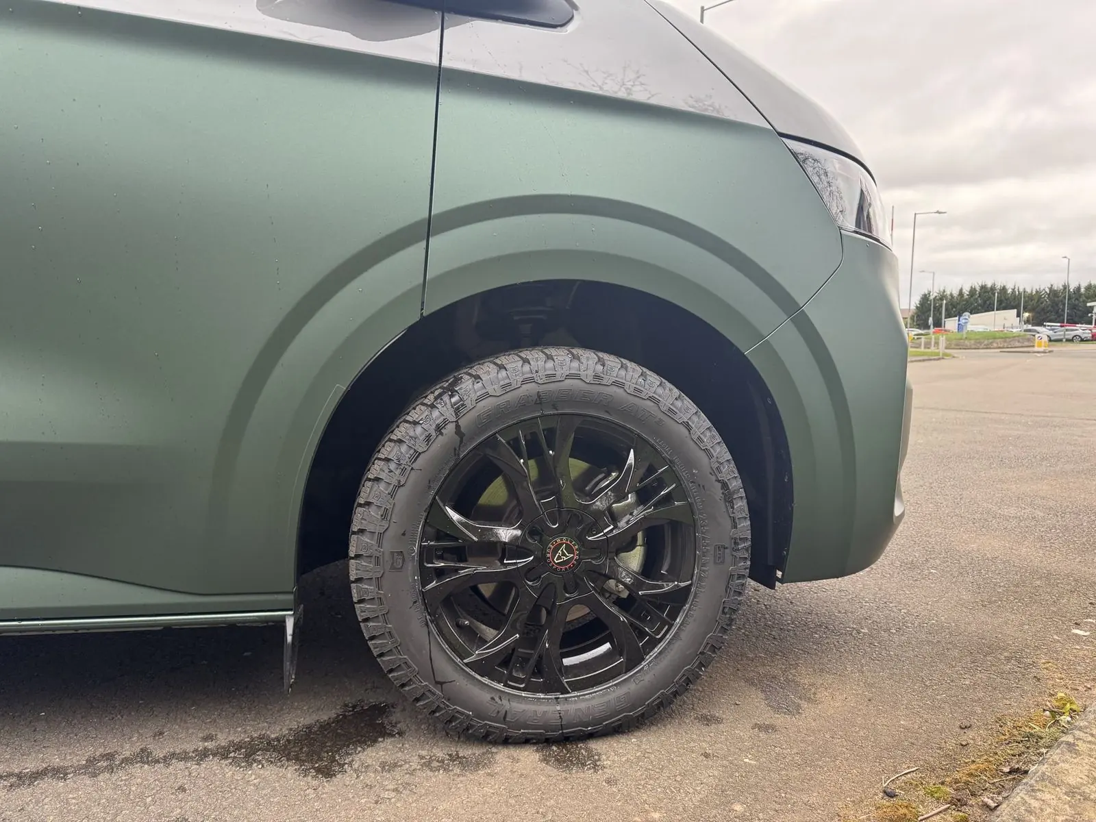 Close-up of the front wheel arch showing matt dark green bodywork, a gloss black multi-spoke alloy wheel and a General Grabber AT3 all-terrain tyre.