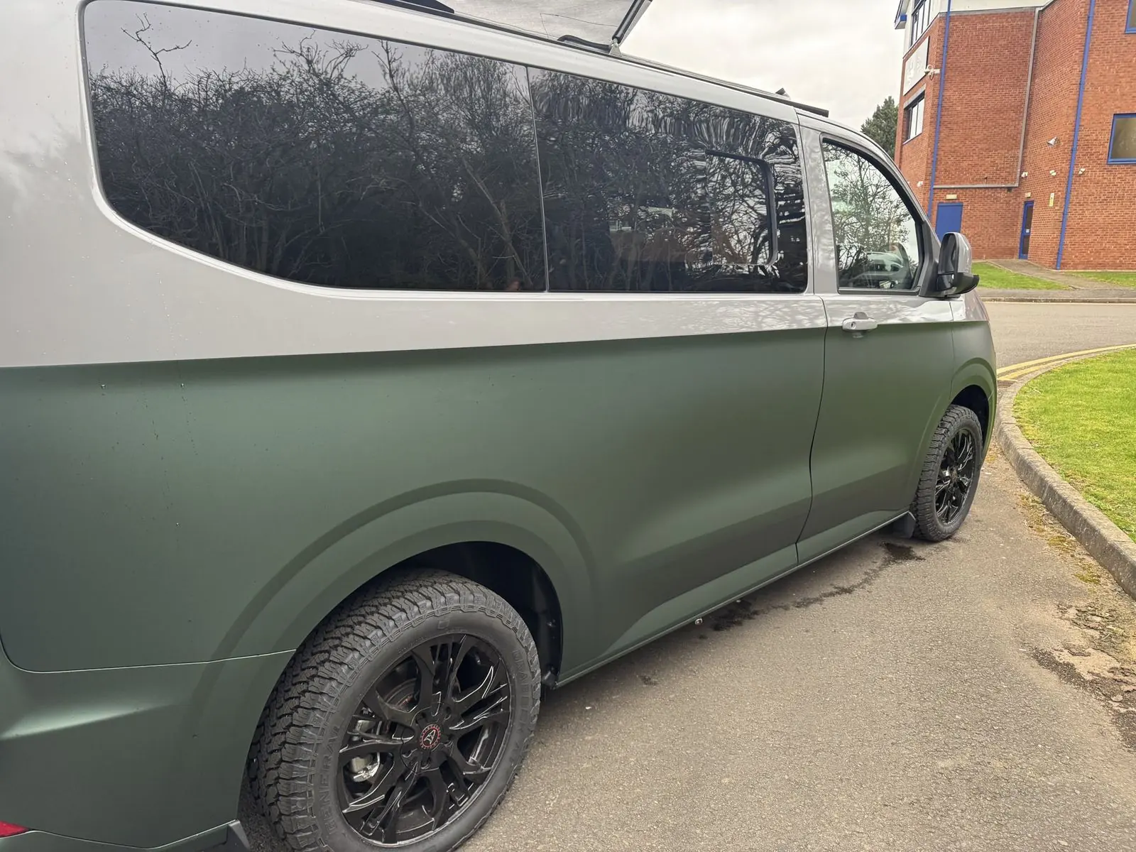Rear three-quarter close-up along the side of the van showing the silver upper and matt green lower bodywork, dark tinted windows and black alloy wheels with all-terrain tyres.