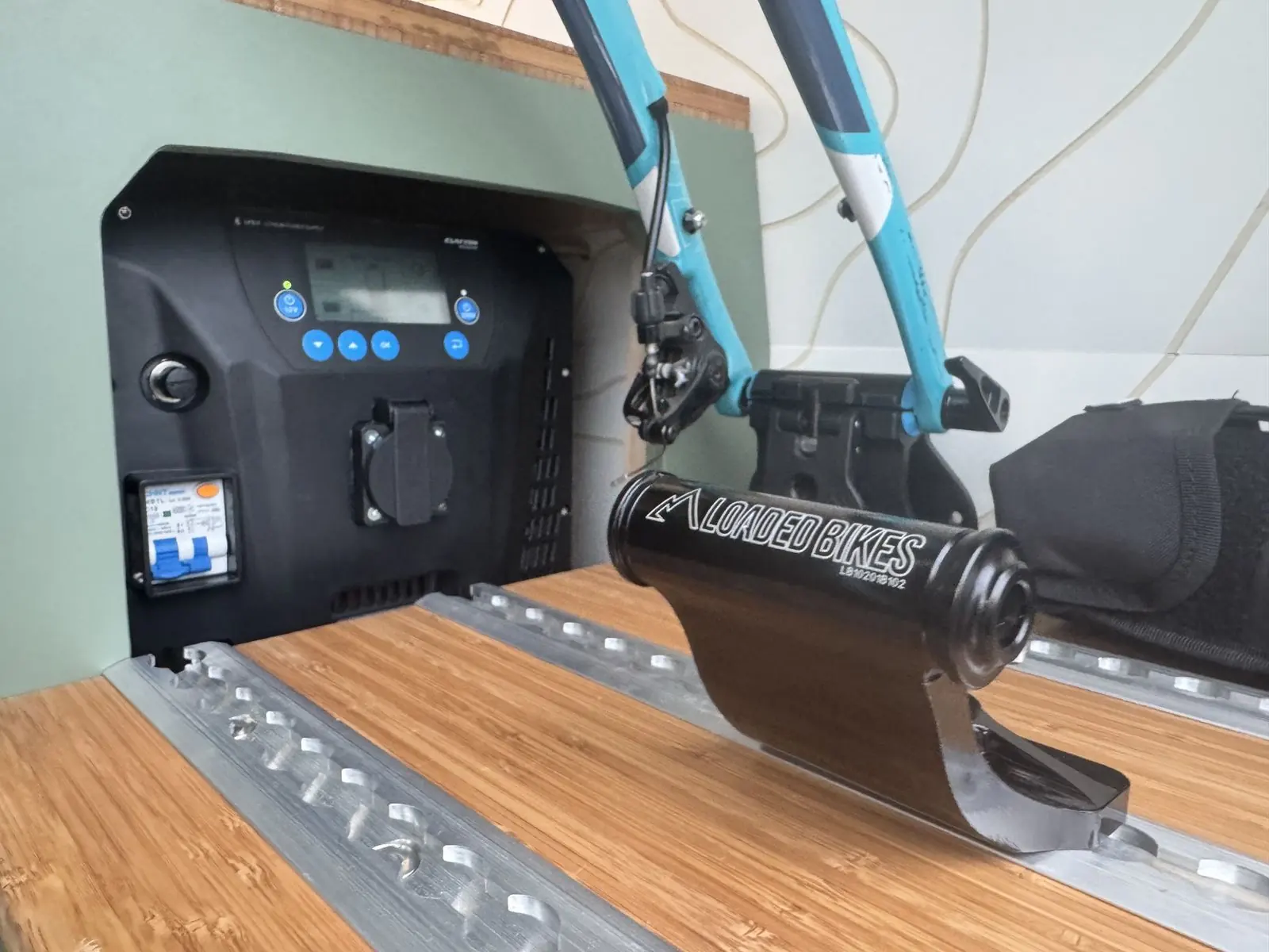 Close-up of a bike fork mount fixed to aluminium rails on the bamboo-topped rear unit, holding a turquoise bike fork, beside a black built-in power unit with a display and socket.