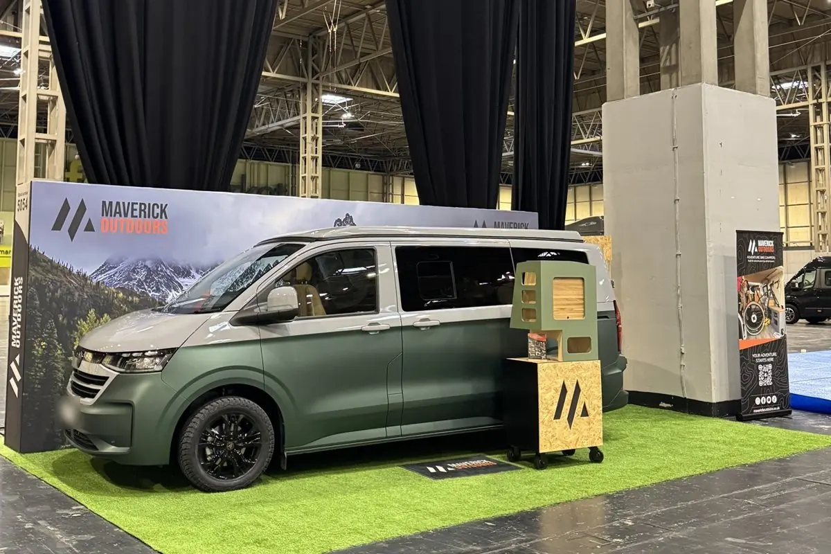 Matt green and silver VW Transporter camper on the Maverick Outdoors stand at the NEC, on green turf in front of a mountain backdrop, with a sage green display unit beside it.