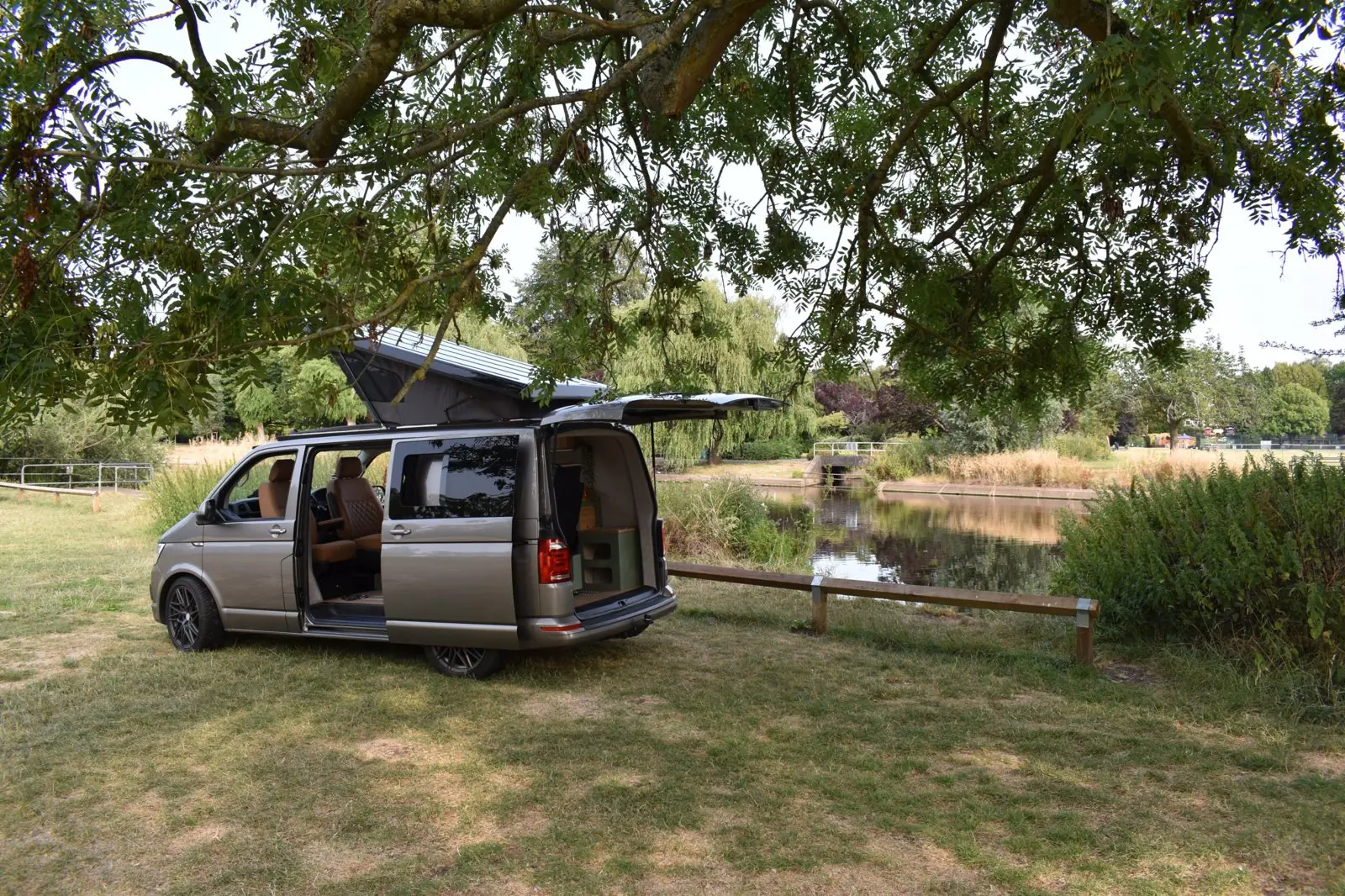 Champagne Volkswagen Transporter T6 campervan parked on grass beside a river under a tree, with its pop-top raised and the side door and tailgate open, showing tan seats and the rear storage unit.