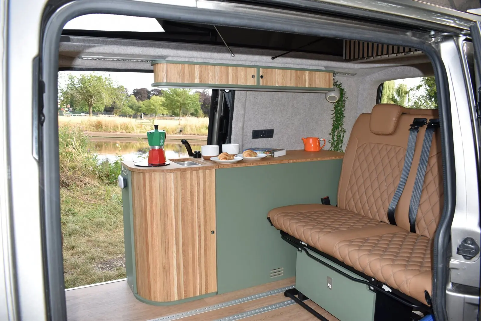 View through the side door of the Brighton interior: a sage green kitchen pod with a curved slatted oak end, bamboo worktop and sink, an oak overhead locker, grey felt lining and a tan diamond-quilted rear bench, with the river beyond.