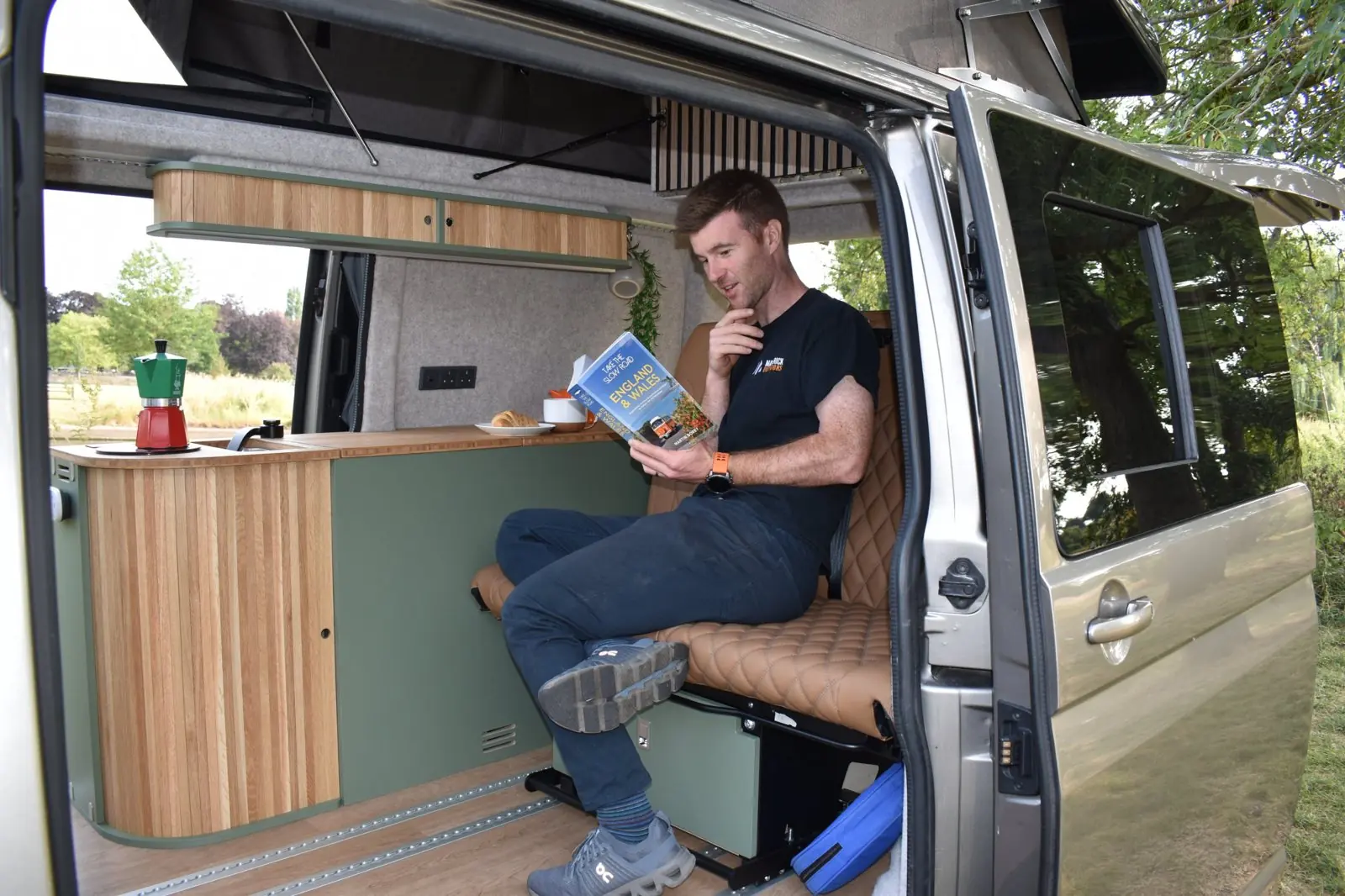 A member of the Maverick team sitting on the tan quilted rear bench reading a road-trip guidebook, with the kitchen pod, bamboo worktop and coffee pot beside him and the pop-top raised above.