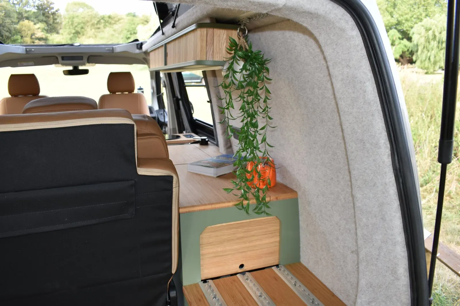 View in through the tailgate: the back of the rear bench, a sage green rear cabinet with a bamboo top and oak door, a slatted bamboo boot floor with rails, felt-lined walls and a hanging plant.