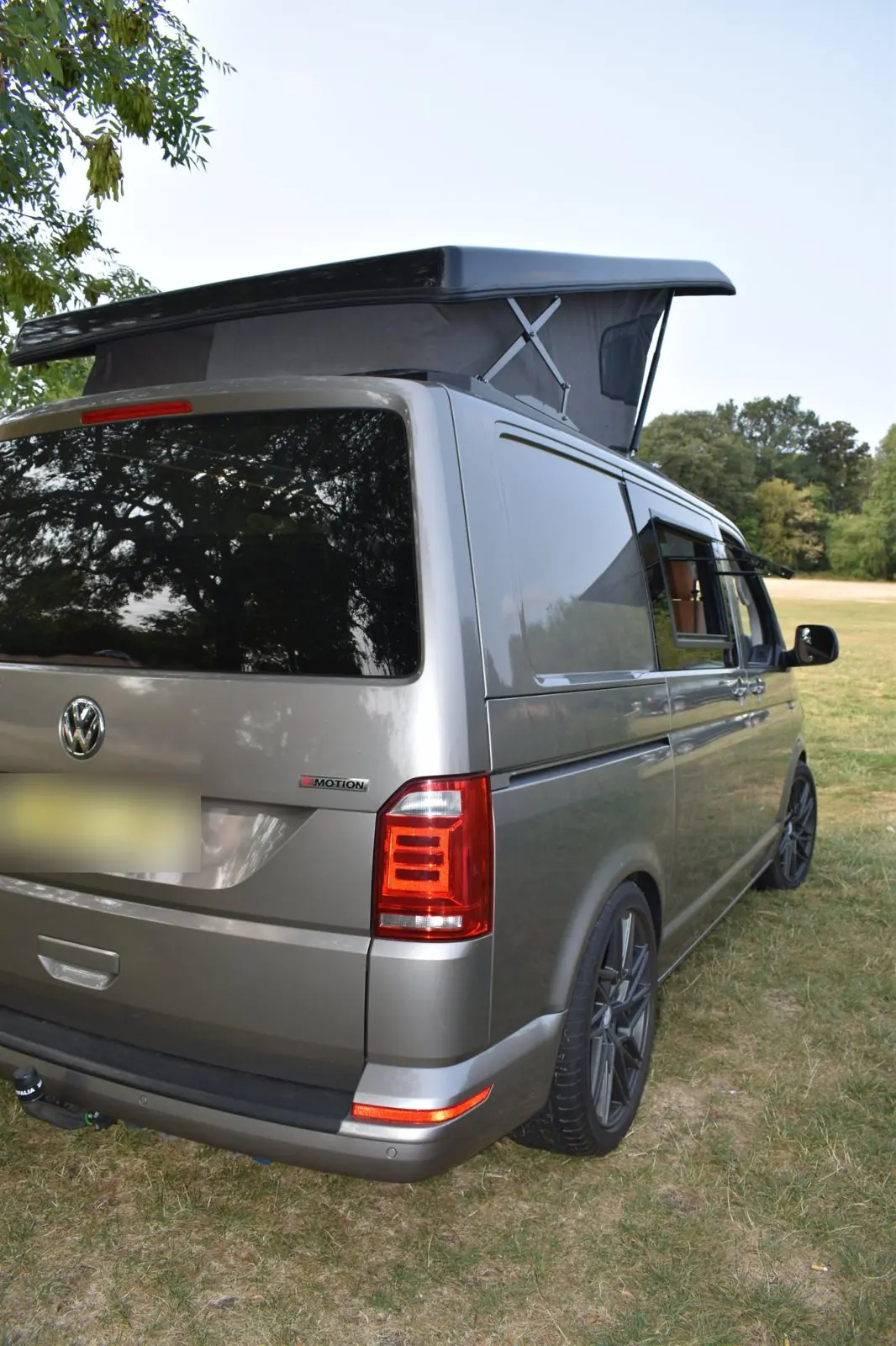 Rear three-quarter view of the champagne Transporter with the dark pop-top raised, a 4Motion badge on the tailgate, an opening side window and dark grey alloy wheels.