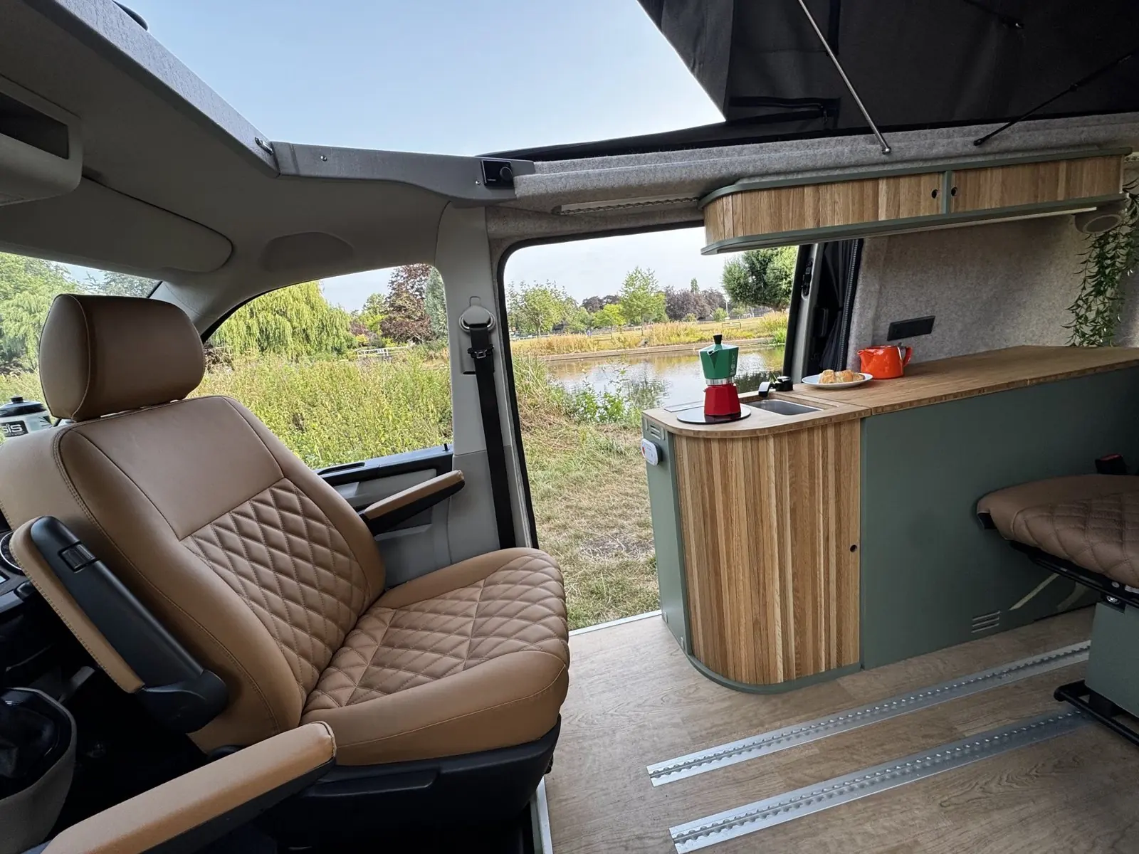 Interior looking across to the open side door and a river beyond: a tan diamond-quilted front seat, a sage green kitchen pod with a curved slatted oak end and bamboo worktop, an oak overhead locker and floor rails.