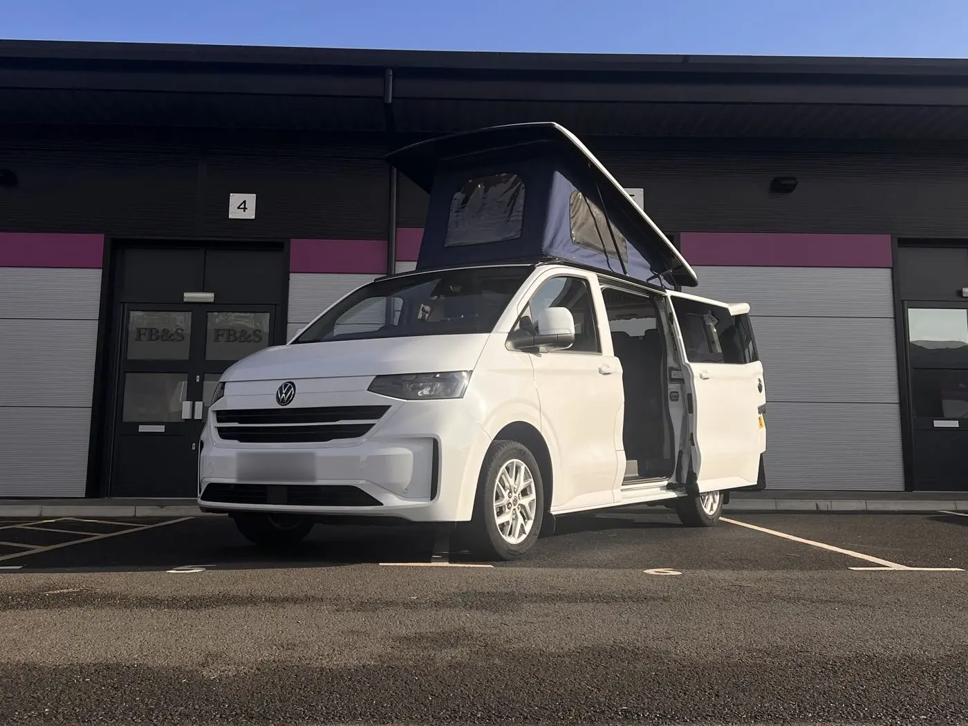 White new-shape Volkswagen Transporter campervan parked outside a row of business units with its navy blue pop-top roof raised and the side door open.