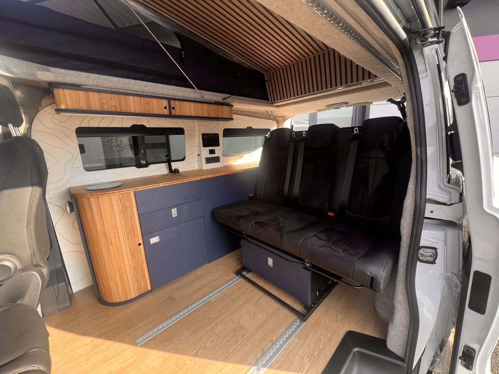 Interior seen through the side door: navy blue kitchen unit with an oak slatted curved end and bamboo worktop, a three-seat rear bench in black, an oak overhead locker, contour-line wall panels and a slatted roof panel.