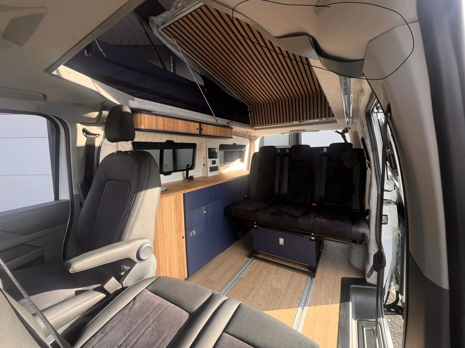 Wide view from the cab across a front seat with armrest to the navy kitchen, bamboo worktop, three-seat rear bench and oak-effect floor with two rails, with the raised roof and slatted panel above.