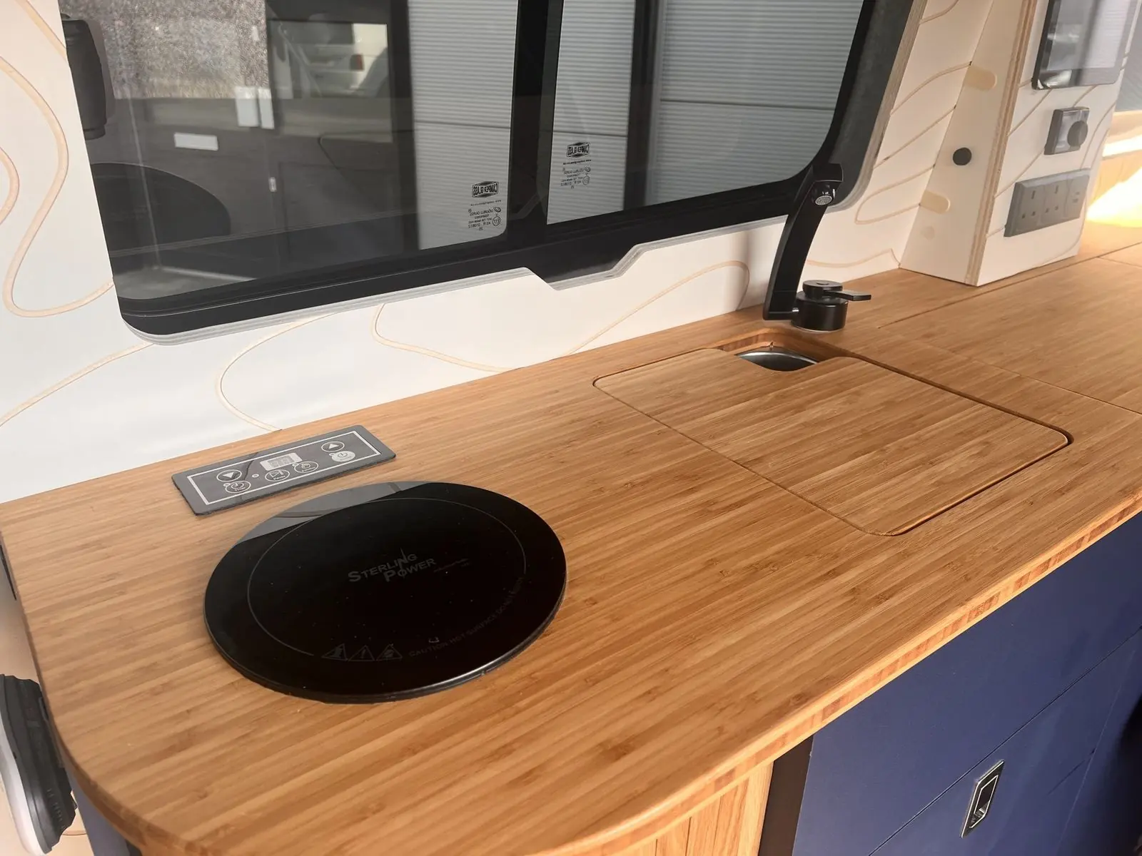 Bamboo worktop with a round black Sterling Power induction hob and touch control panel, a flush bamboo sink cover and a black tap, beneath a sliding side window.