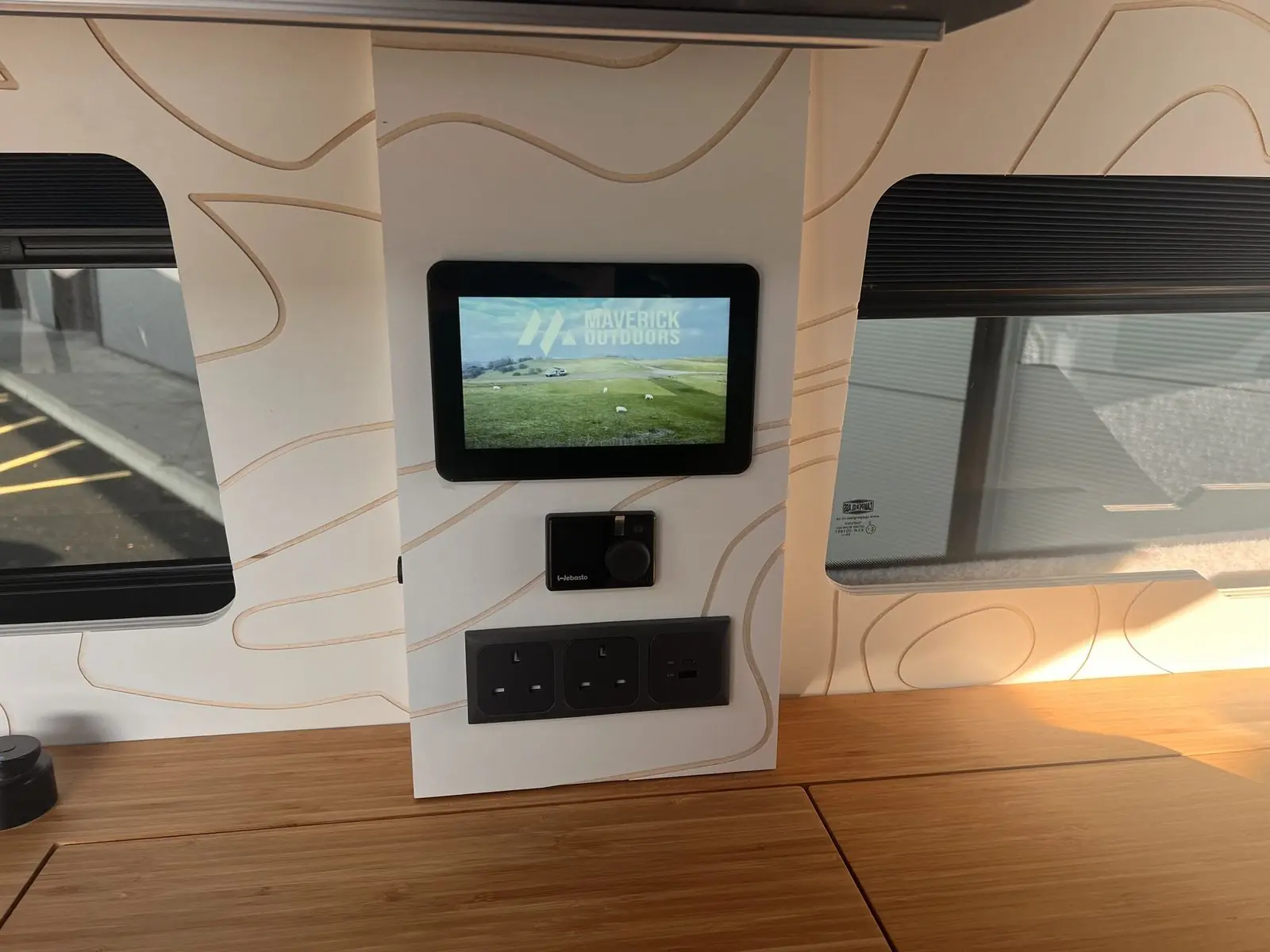 Wall panel engraved with a contour-line pattern carrying a touchscreen showing a Maverick Outdoors image, a Webasto dial controller and a triple socket panel, between two windows with blinds.
