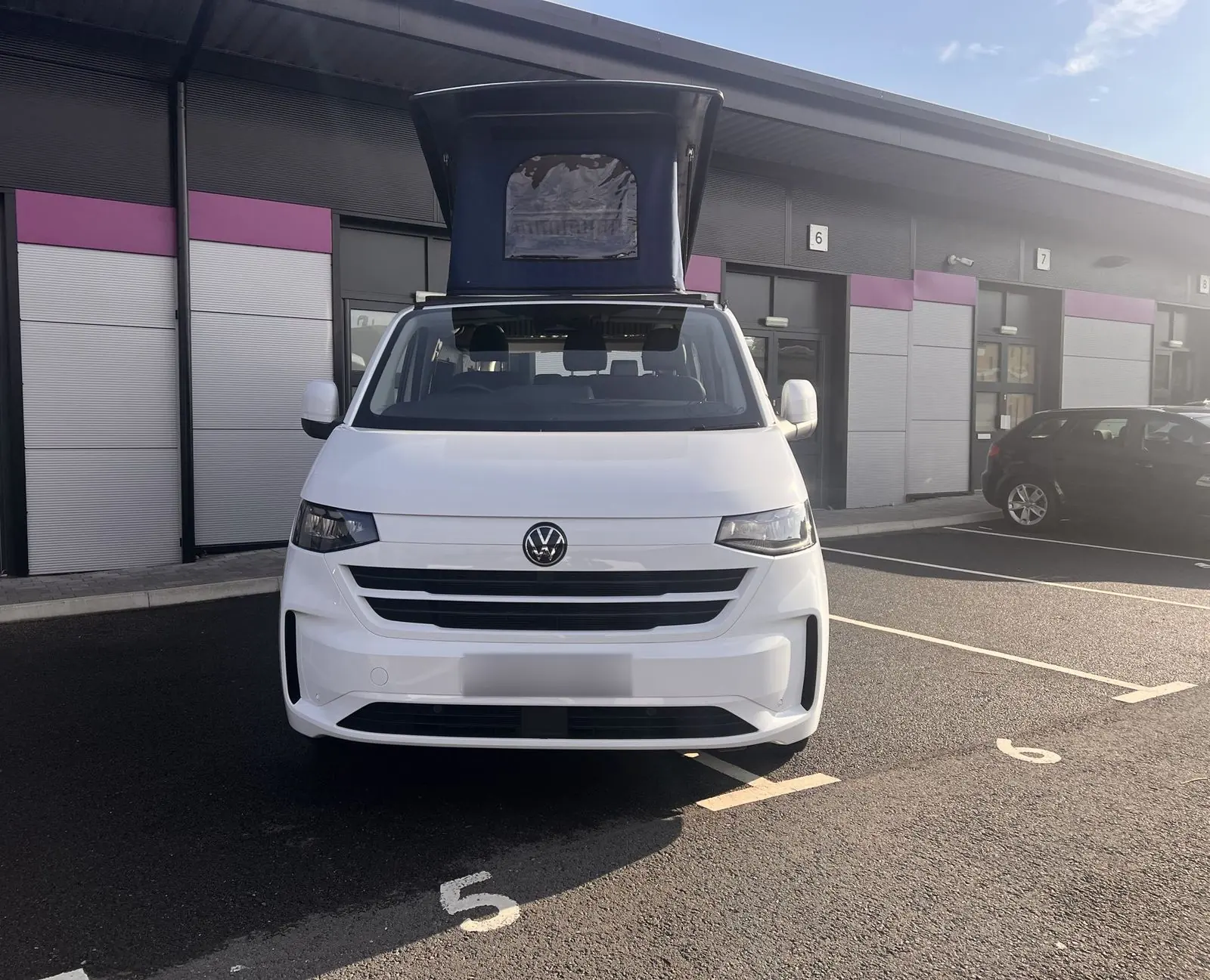 Front view of the white Volkswagen Transporter campervan with its navy pop-top raised, showing the clear front window panel in the canvas.