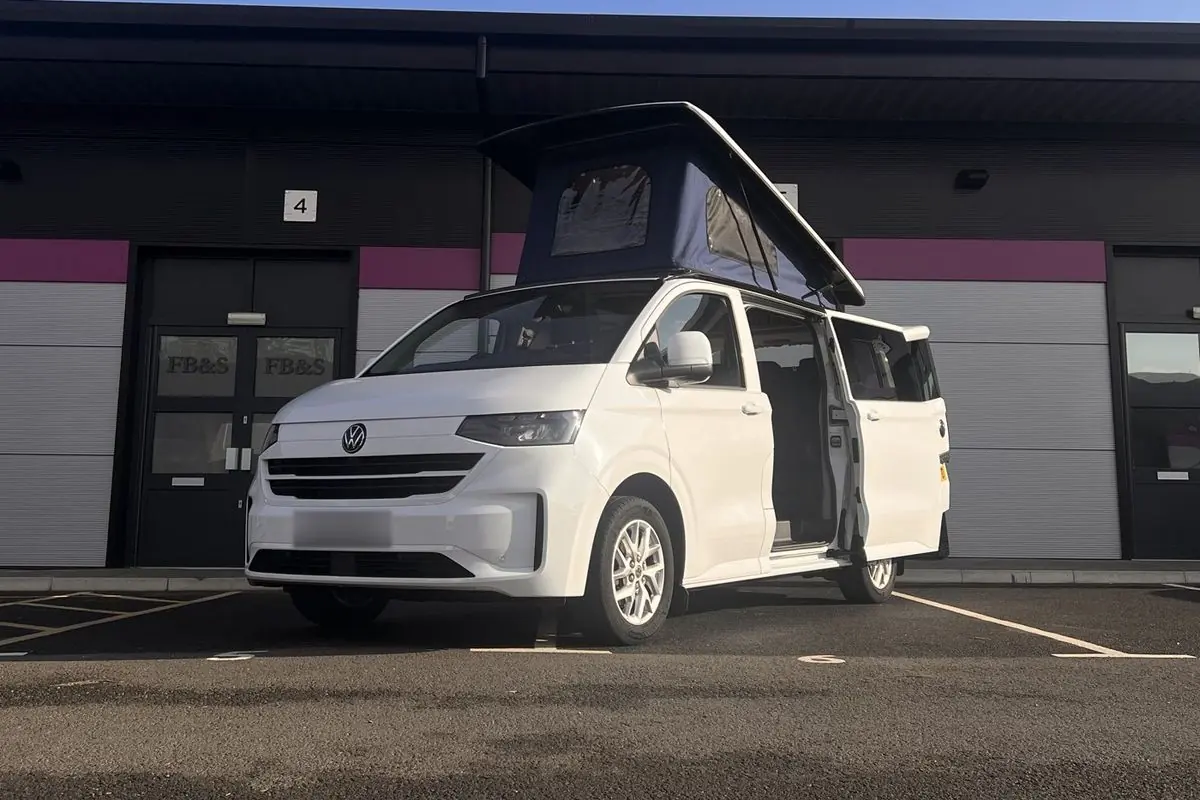 White new-shape Volkswagen Transporter campervan parked outside a row of business units with its navy blue pop-top roof raised and the side door open.