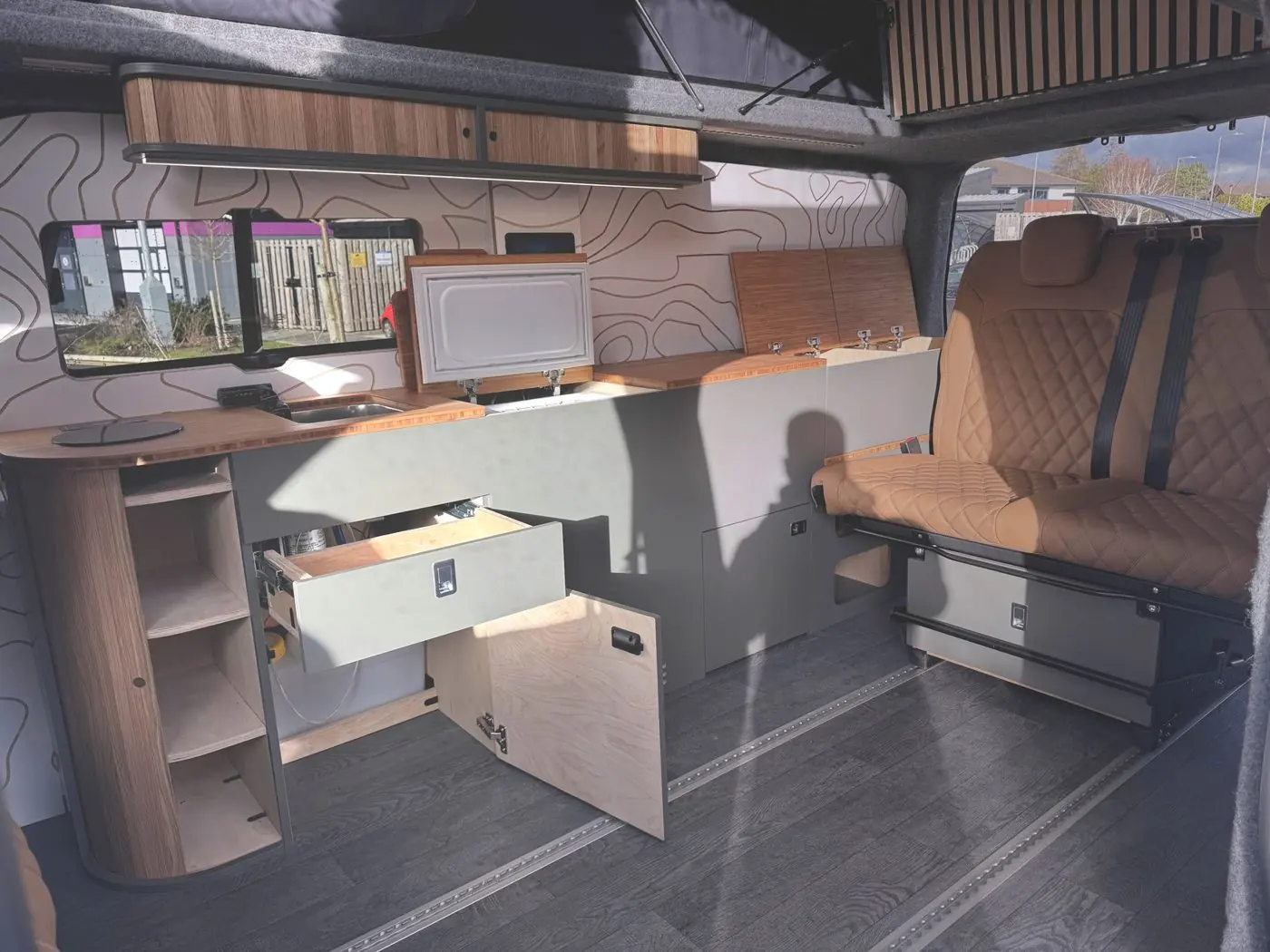 Camper interior with the kitchen drawer and cupboard open, tan rear seat and grey floor with rails
