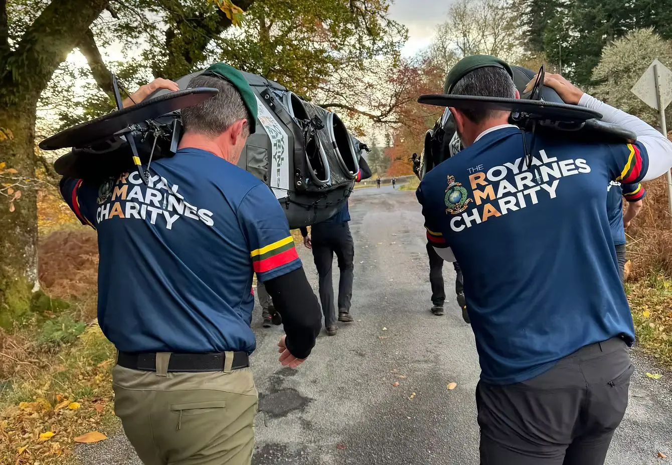 Two people in Royal Marines Charity shirts carrying kayaks along a wooded lane