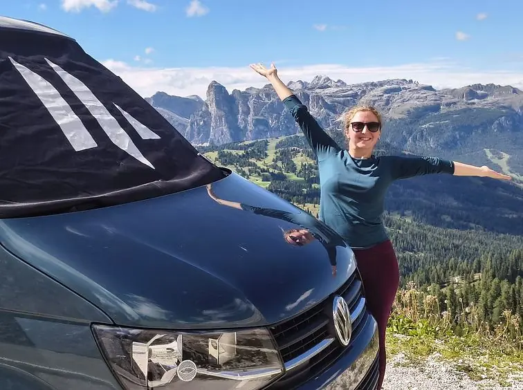 A Maverick owner with arms outstretched beside her van, mountains behind
