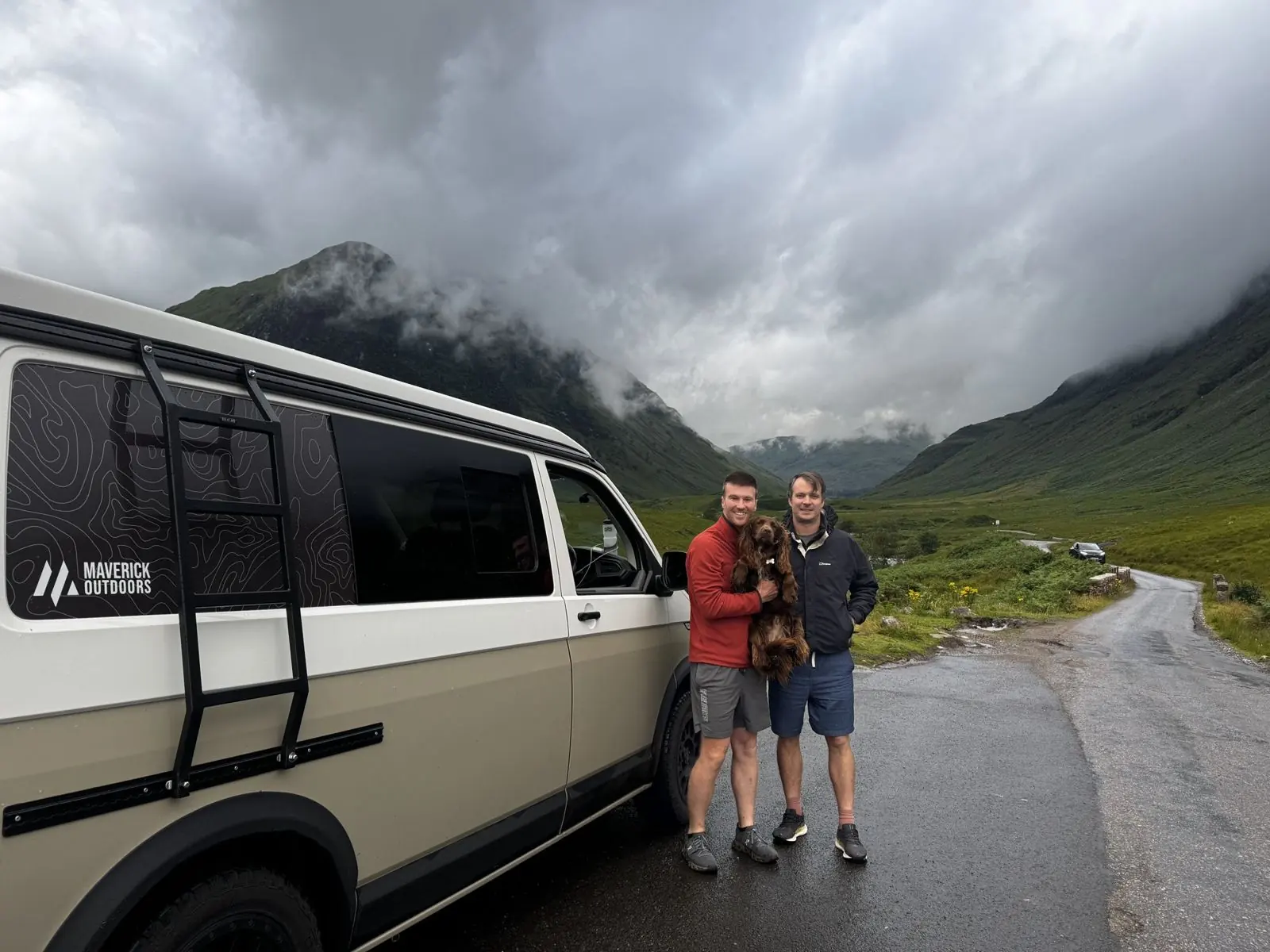 Two of the Maverick team with their spaniel beside a Maverick Outdoors camper in a Highland glen